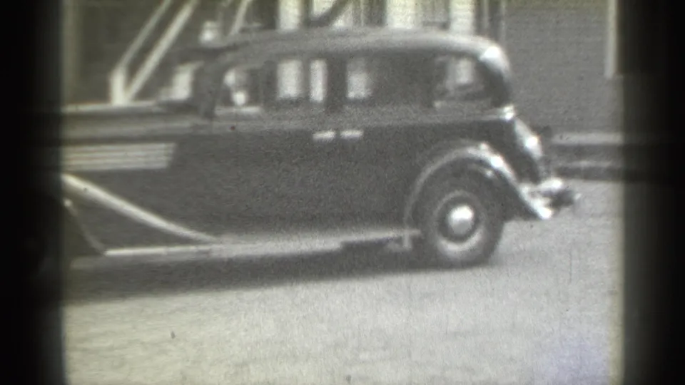 1939 New York home-movie frame focused on the rear portion of a dark sedan, with stairs, windows, and soft film grain visible.