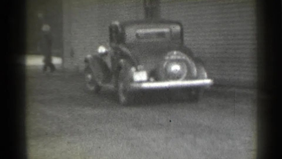 1939 New York street frame with a dark pre-war car and a distant person in dark clothing near a building entrance.