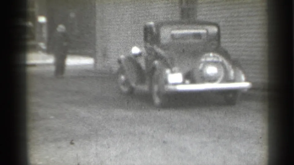 Rear view of a dark pre-war sedan on a New York street, with a blurred pedestrian near the edge of the frame.