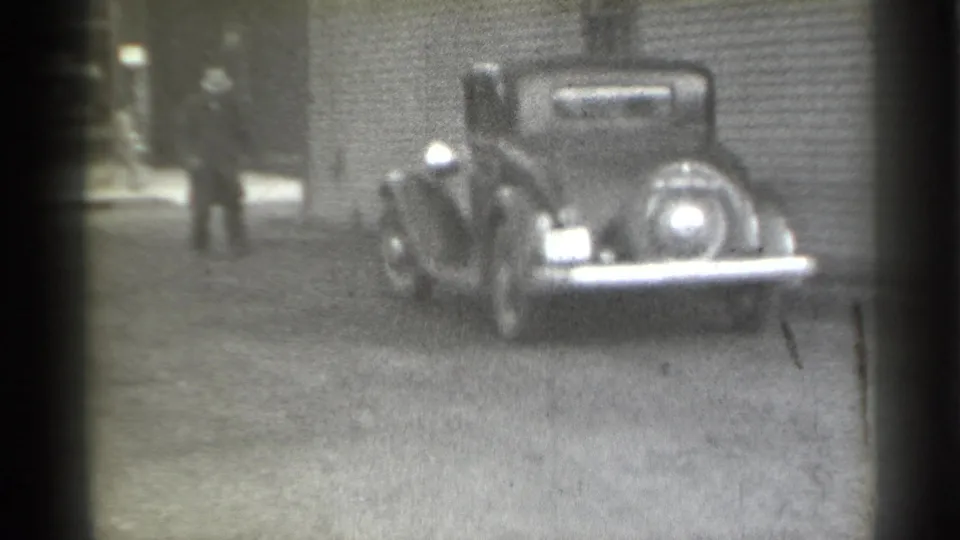 1939 New York 8mm frame showing a dark pre-war car parked near a building as pedestrians move behind it.