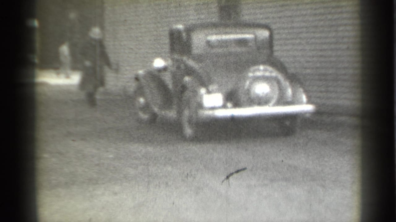 Suited man walking near a parked vintage car on a dirt lot.