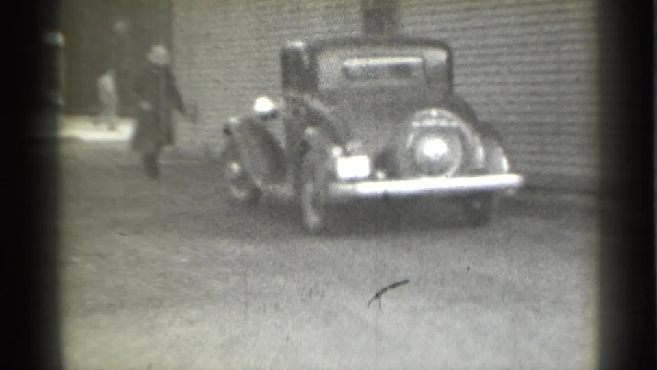 1939 New York home-movie frame showing a suited man walking near a parked vintage car beside a corrugated building.