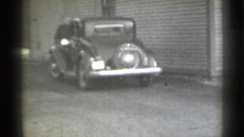 1939 New York frame with a man partly obscured beside a parked dark automobile in front of a corrugated metal facade.