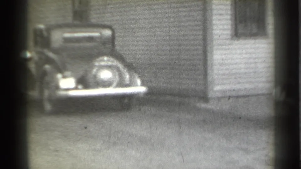 1939 New York home-movie view of a parked vintage car with a rear spare tire near a corrugated building and doorway.