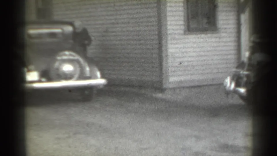 1939 New York frame showing the parked vintage car beside a corrugated building, with another vehicle edge entering at right.