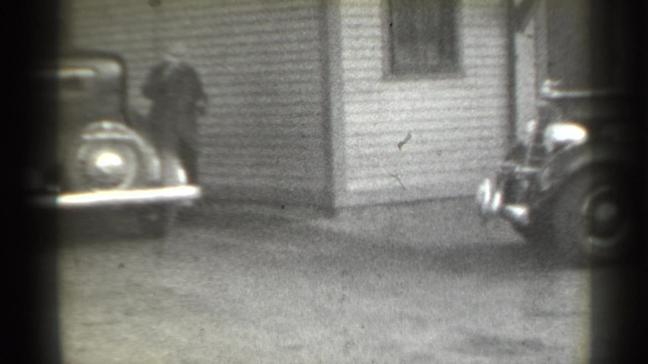 Man near a clapboard house with part of a vintage car at the edge of the frame.