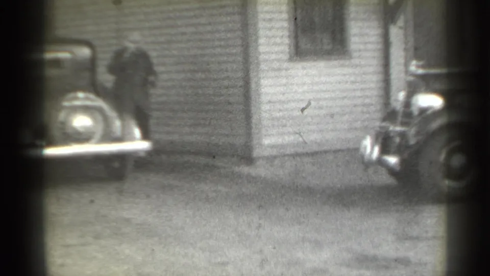 In 1939 New York home-movie footage, a man in a coat and hat stands near a clapboard house as a parked car partly enters the frame.