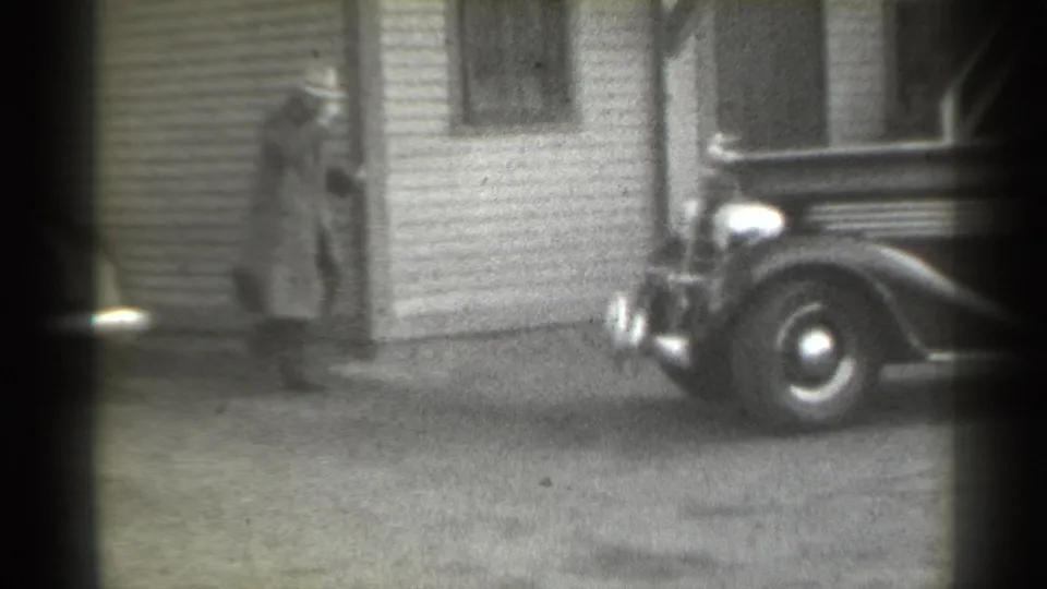 A man in a long coat and hat stands near a clapboard house doorway while the front of a 1930s-style car is parked close by.