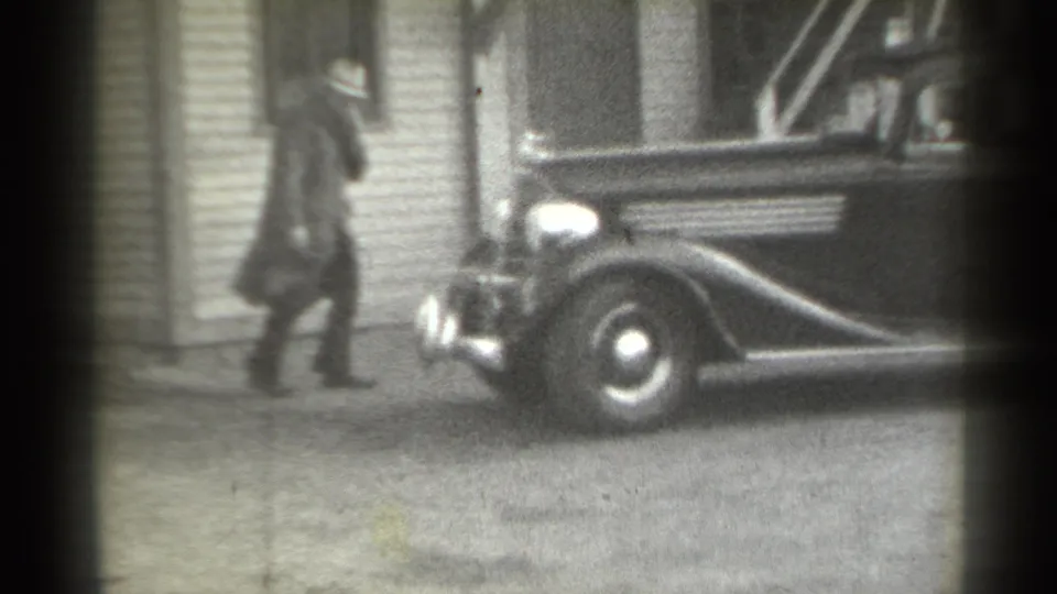 The man walks along the house exterior past the front fender and wheel of a parked vintage automobile in black-and-white 8mm footage.