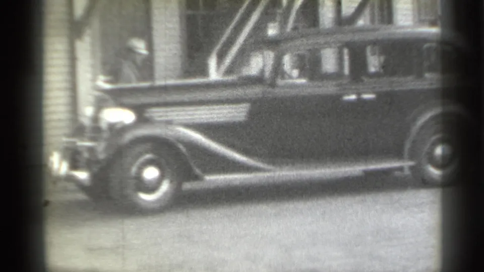 A parked 1930s-style automobile is seen in side view beside a clapboard house, with part of a doorway and window behind it.
