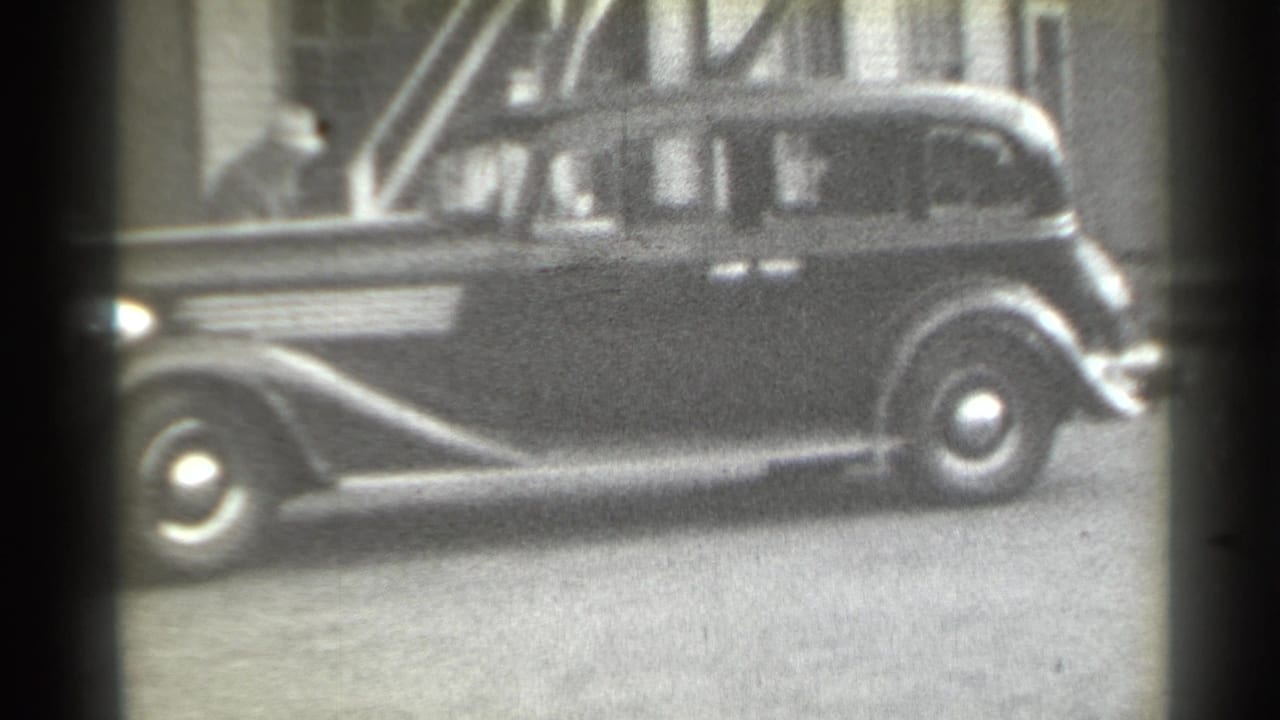 1939 New York home-movie frame showing a dark sedan in profile on a city street, with a hat-wearing person partly visible near the building entrance.