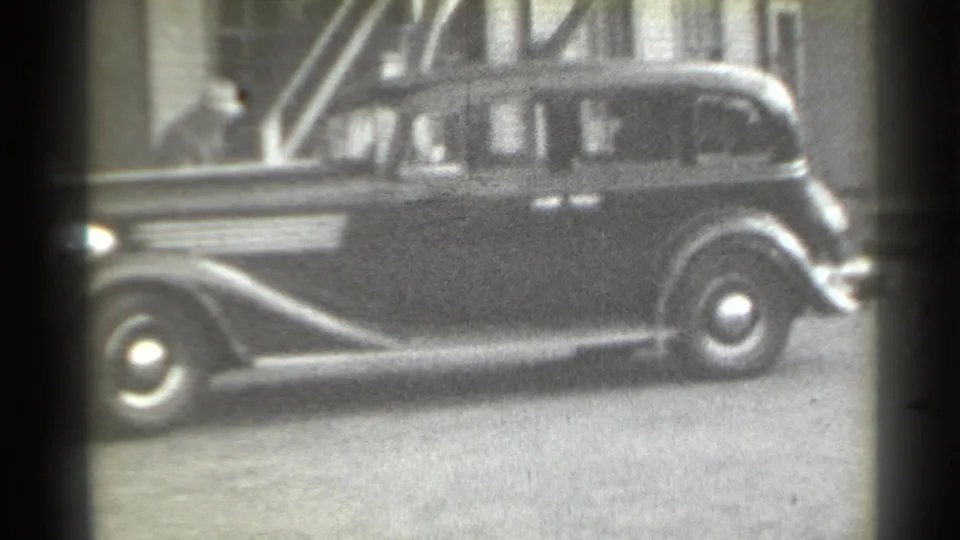 1939 New York home-movie frame showing a dark sedan in profile on a city street, with a hat-wearing person partly visible near the building entrance.