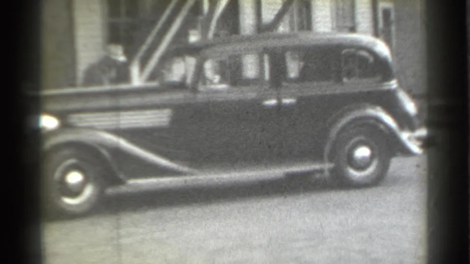 1939 New York street frame showing the side of a parked dark sedan, with masonry architecture and a partly obscured person in the background.
