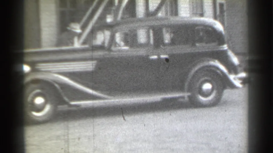 1939 New York home-movie frame showing a parked sedan with soft silhouettes inside or near the car and a building facade behind it.