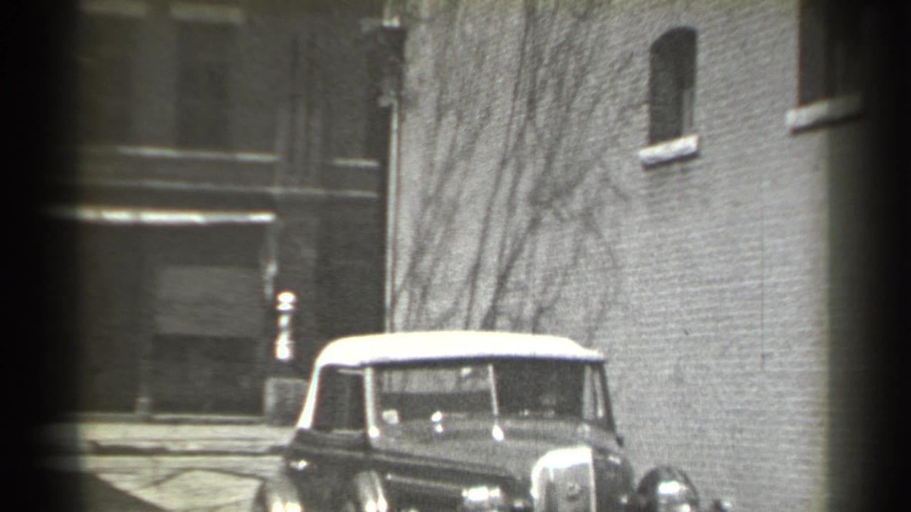 1939 New York home-movie frame showing a vintage convertible with a pale soft top near brick buildings and winter-bare branches.