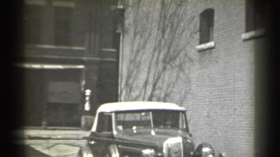 1939 New York home-movie frame showing a vintage convertible with a pale soft top near brick buildings and winter-bare branches.