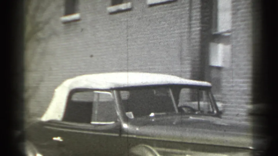 Side view of the vintage convertible in 1939 New York, its pale soft top and chrome trim visible against a brick building wall.