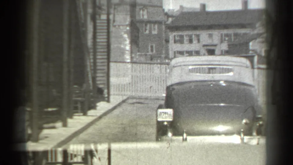 A dark convertible approaches on a quiet New York residential street in 1939, with houses and wooden fencing behind it.