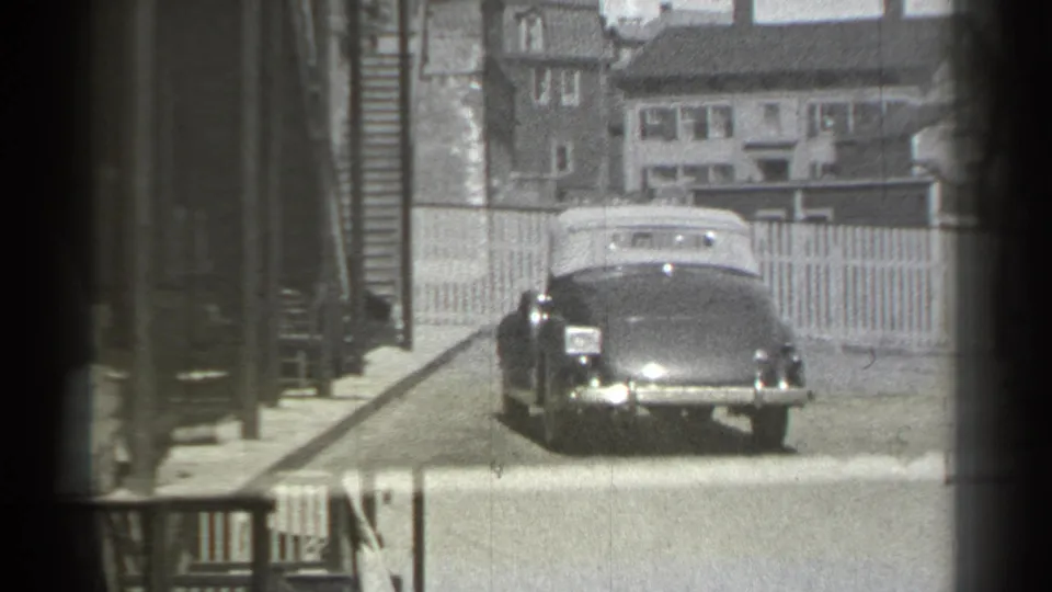The convertible moves along the residential road, framed by fences, houses, utility lines, and the dark edges of the 8mm frame.