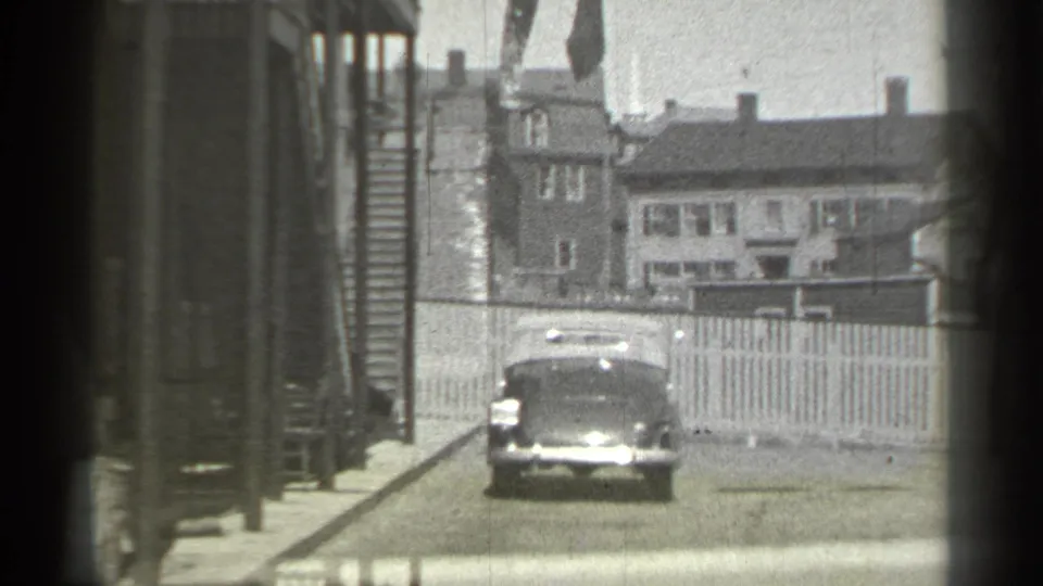 The car faces the camera near the center of the street, with detached homes, fencing, and soft black-and-white film texture visible.