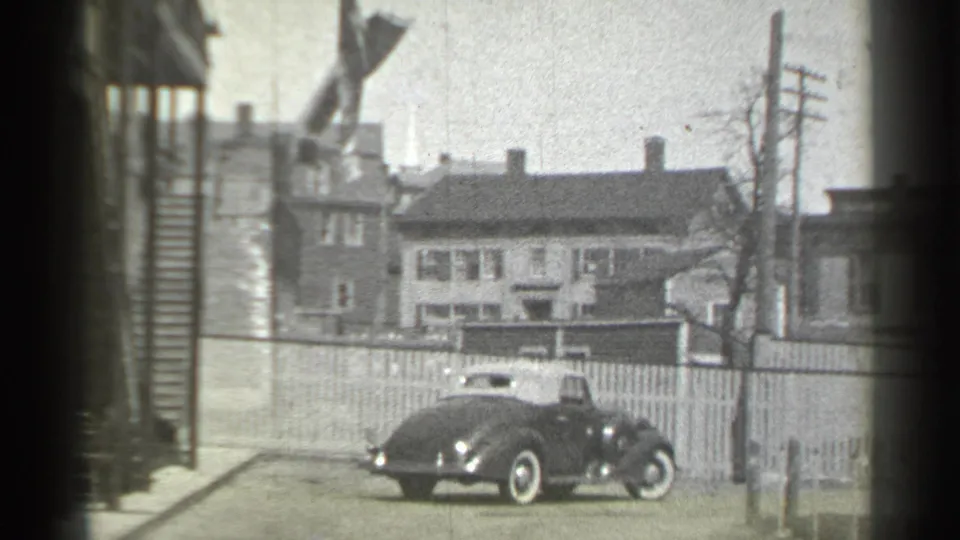 The dark convertible turns across the New York residential street, showing its side profile, light top, whitewall tires, and nearby houses.