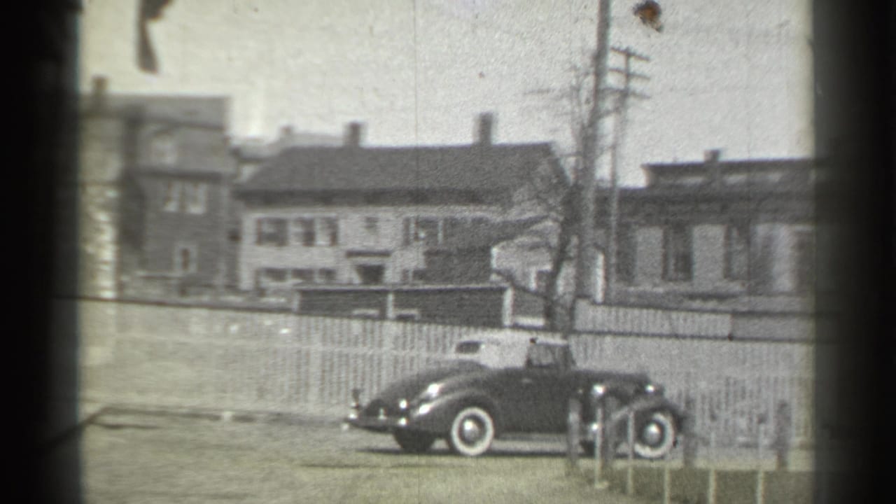 1939 New York home-movie frame showing a dark classic car near a driveway, with row houses, utility poles, and fencing behind it.