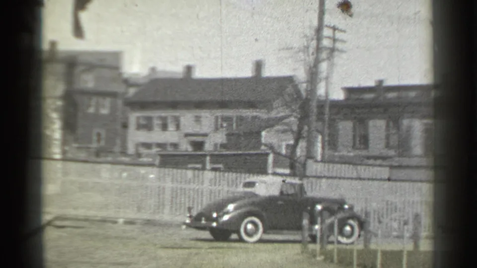 1939 New York home-movie frame showing a dark classic car near a driveway, with row houses, utility poles, and fencing behind it.