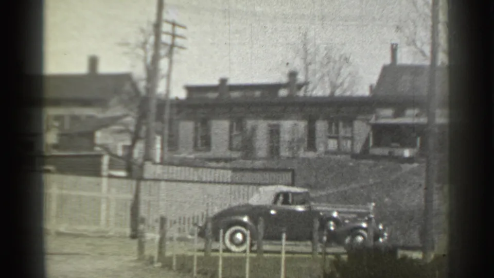 1939 New York frame with the car farther along the street, seen against a fence, bare trees, utility poles, and residential buildings.