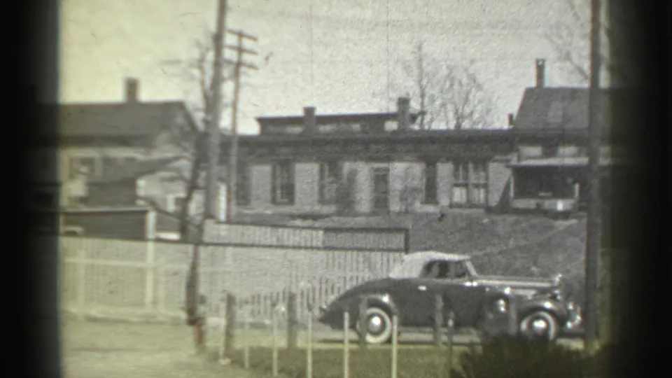 1939 New York home-movie view of a dark rounded automobile beside a fence, with chimneys, houses, and utility poles behind it.