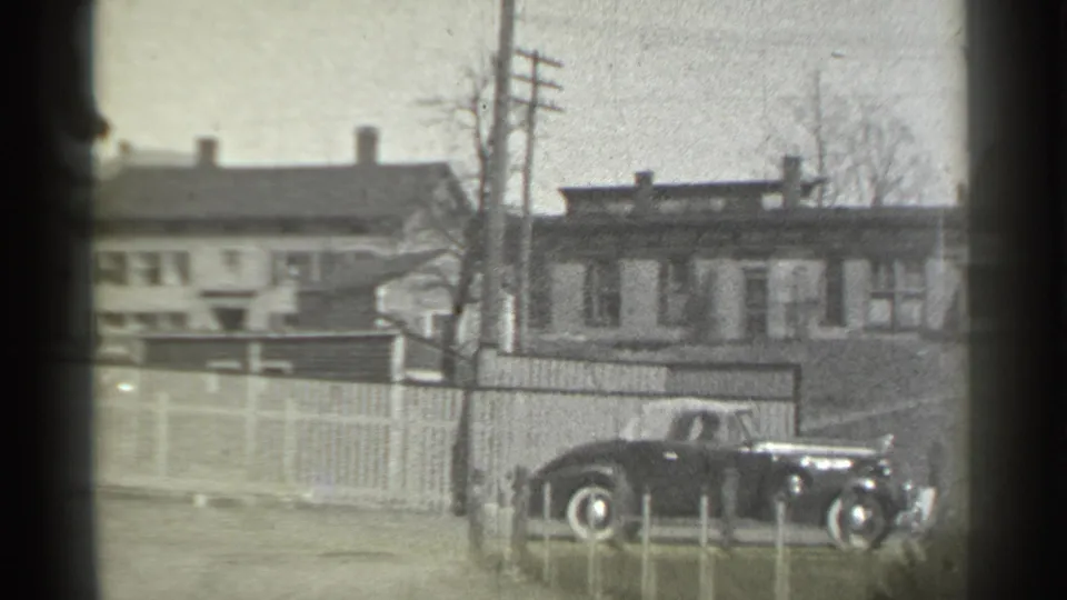 1939 New York frame showing the classic car near the edge of the shot, framed by fencing, bare trees, row houses, and dark gate shadows.