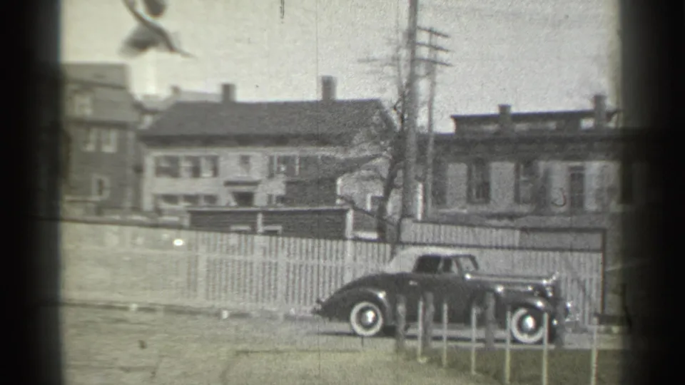 1939 New York home-movie frame showing a dark vintage car near houses, a picket fence and utility poles.