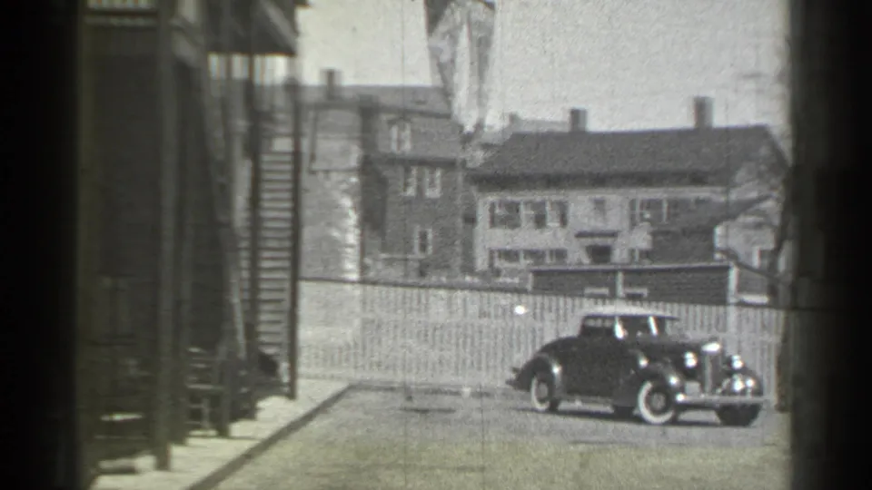 1939 New York frame with a dark automobile reversing near a fence, houses and a sidewalk edge.