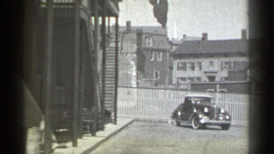 Black-and-white 1939 New York frame showing the rear of a vintage car near houses, fencing and a sidewalk.