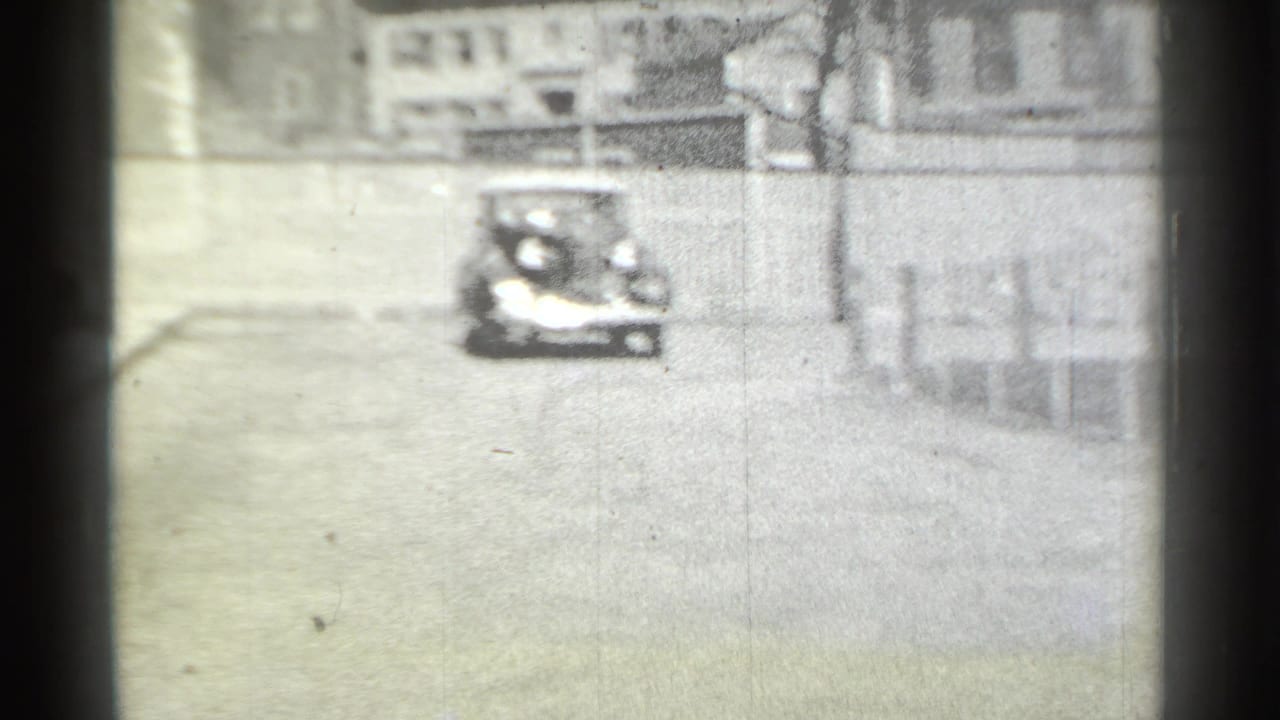 In 1939 New York home-movie footage, a distant classic car approaches on a quiet residential street lined with fences and homes.