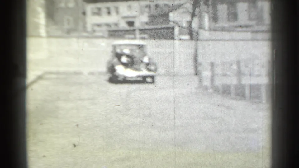 In 1939 New York home-movie footage, a distant classic car approaches on a quiet residential street lined with fences and homes.
