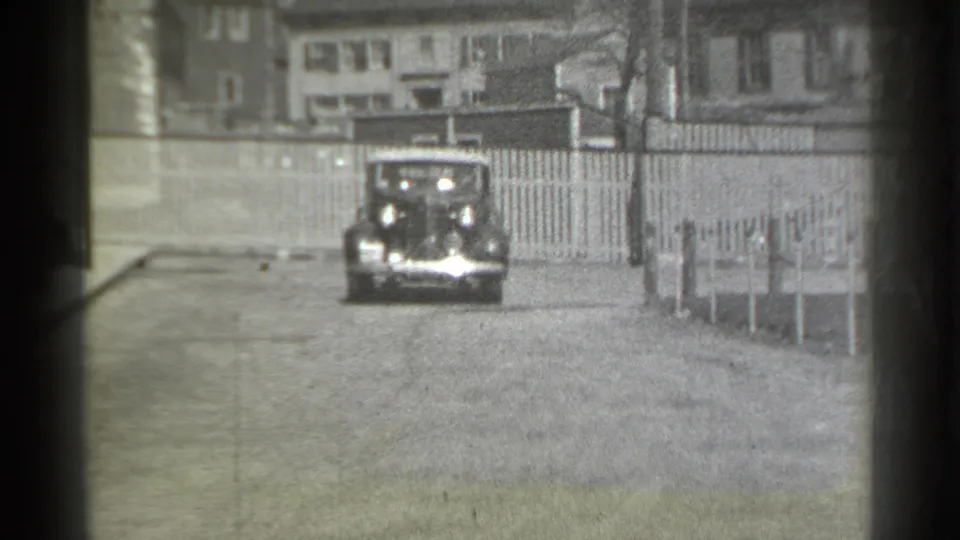 The classic car moves closer toward the camera on a New York residential street, with fencing and multi-story houses behind it.