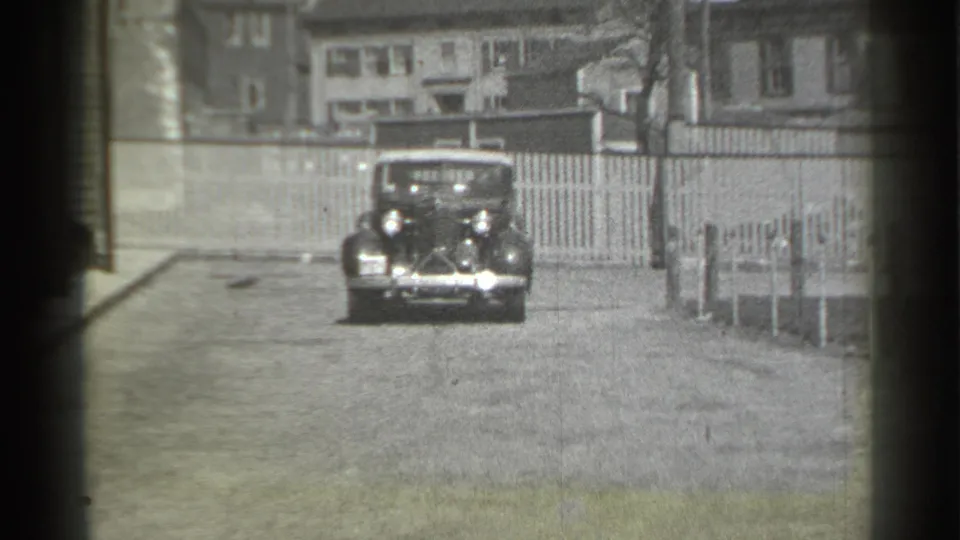 A front-facing classic car approaches the stationary 8mm camera on a quiet 1939 New York residential block.