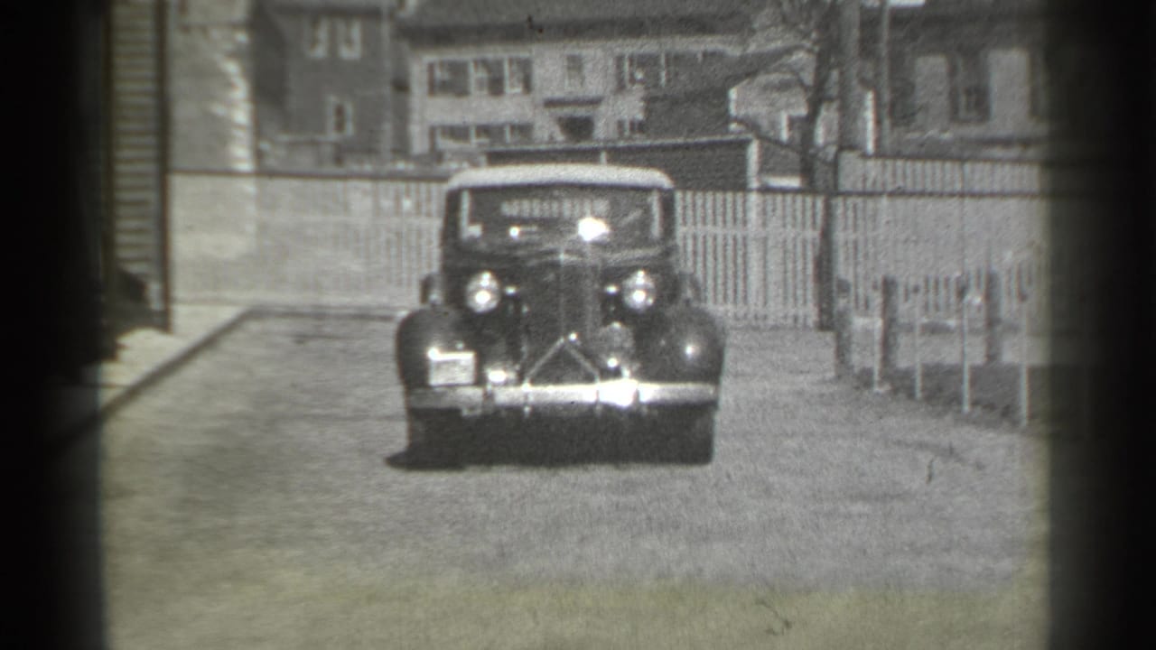 Vintage convertible approaches on a residential street with houses and a picket fence behind it.