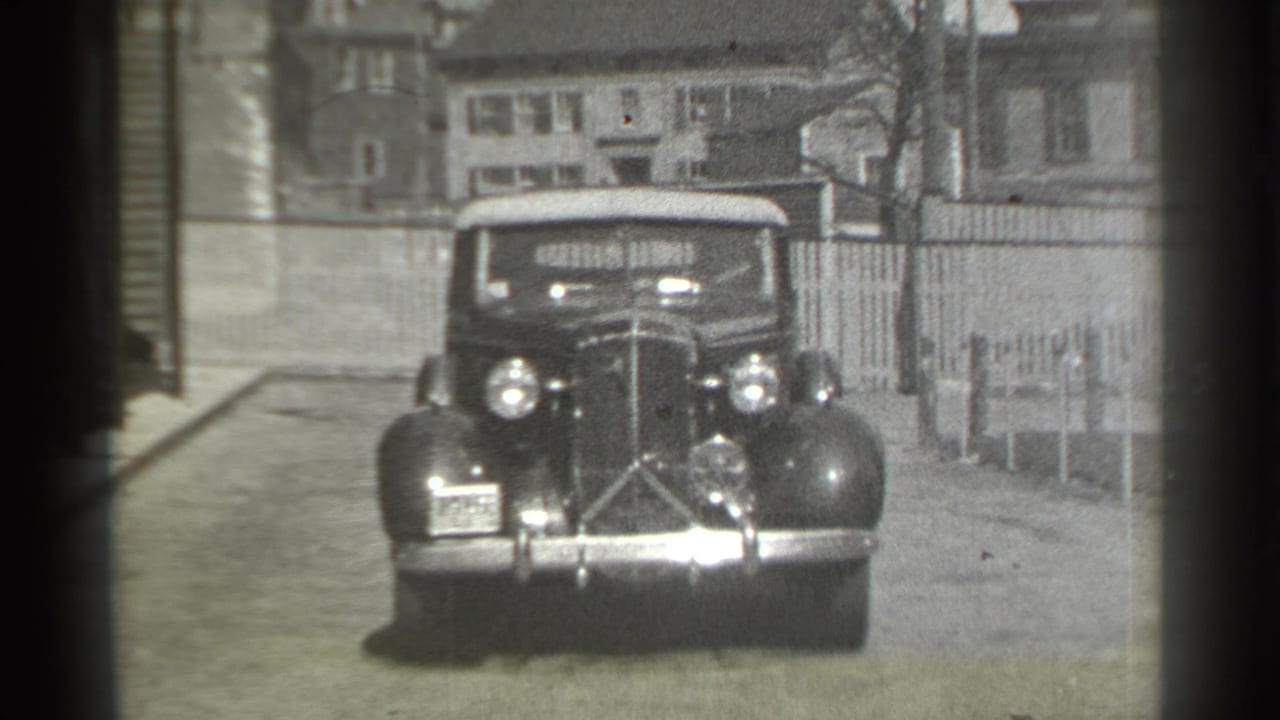 Convertible drives closer toward the camera with headlights visible on a residential street.