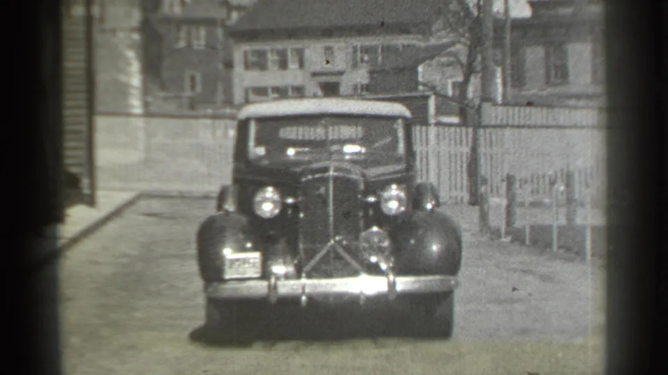 The vintage convertible nears the camera on a New York residential street, framed by houses, fencing, and 8mm film edges.