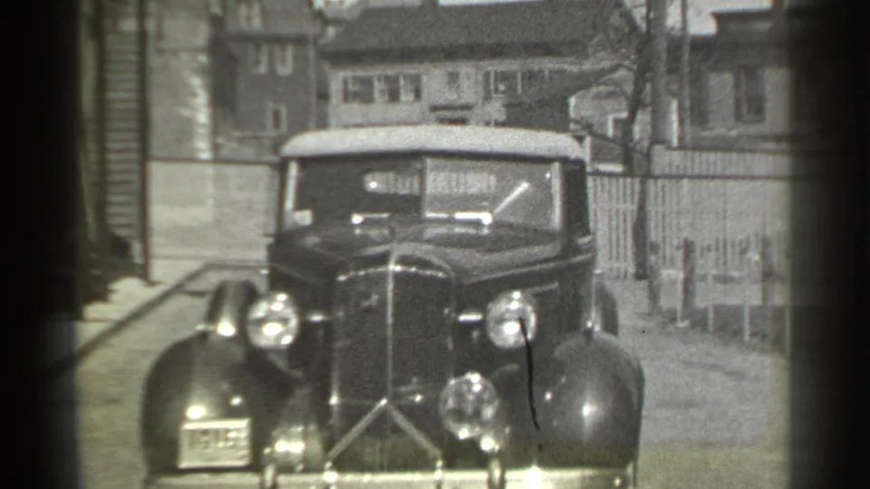 Close 1939 New York frame of the convertible approaching the corner, with its grille, headlamps, soft top, fence, and houses visible.