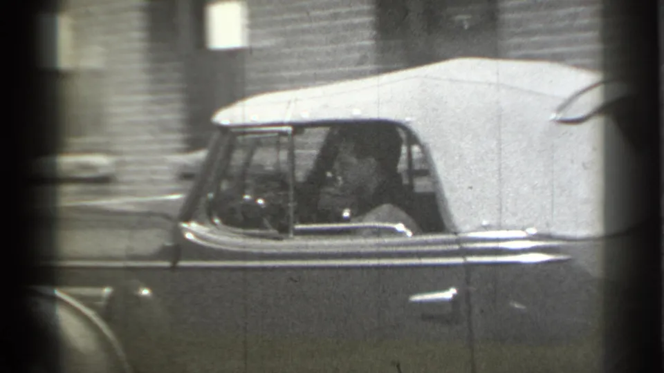 A man is visible through the rolled-down side window of the 1930s convertible as it moves through a New York street.