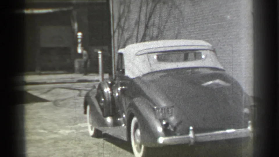 The 1930s convertible is viewed from behind after the turn, with its white soft top, rounded trunk, and urban street surroundings visible.