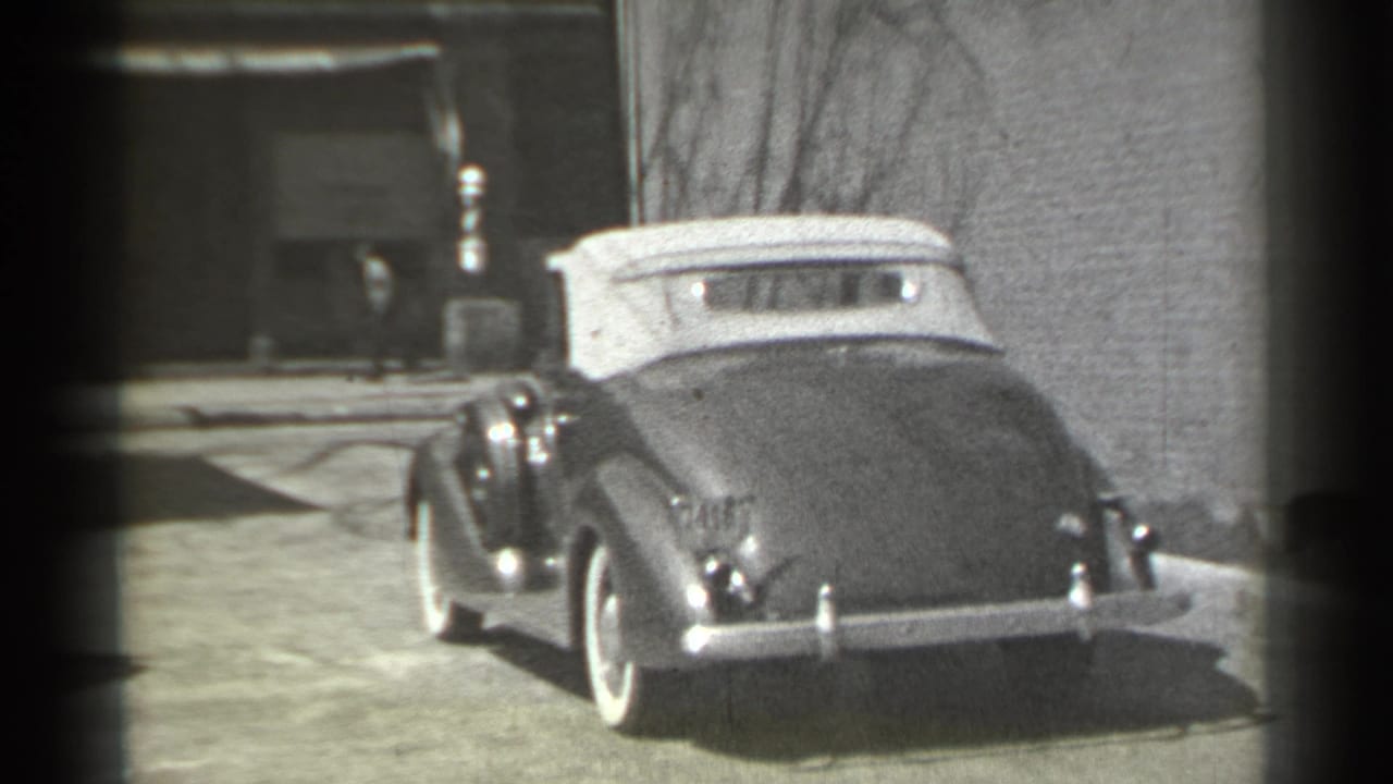 Dark convertible with a light top driving along a quiet street beside a building wall.