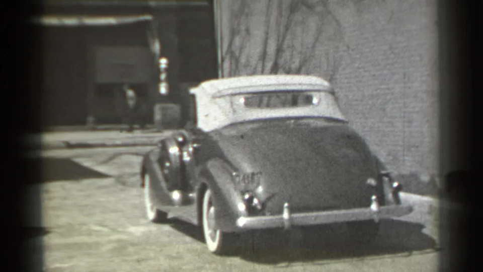 1939 New York home-movie frame showing a dark convertible with a light top passing a masonry building and curb.