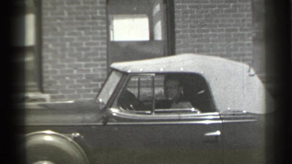 1939 New York 8mm frame showing a classic convertible parked beside brick buildings, with a driver visible through the side window.