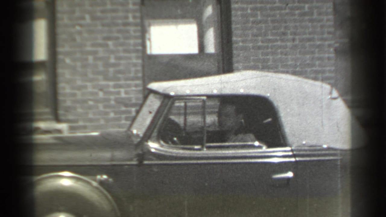 Man seated inside a vintage convertible beside a brick building.