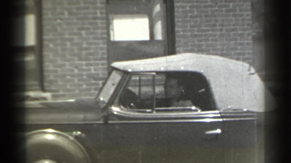 In 1939 New York home-movie footage, a man sits at the wheel of a vintage convertible with a light soft top near a brick facade.