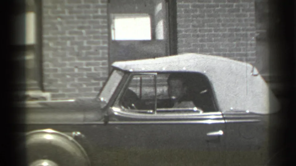 A man is visible through the side window of a light-topped convertible parked in front of a brick building in New York, 1939.