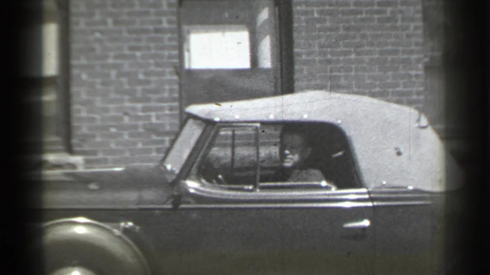 The driver appears more clearly in the open side area of the vintage convertible, with brickwork and a doorway behind the car.